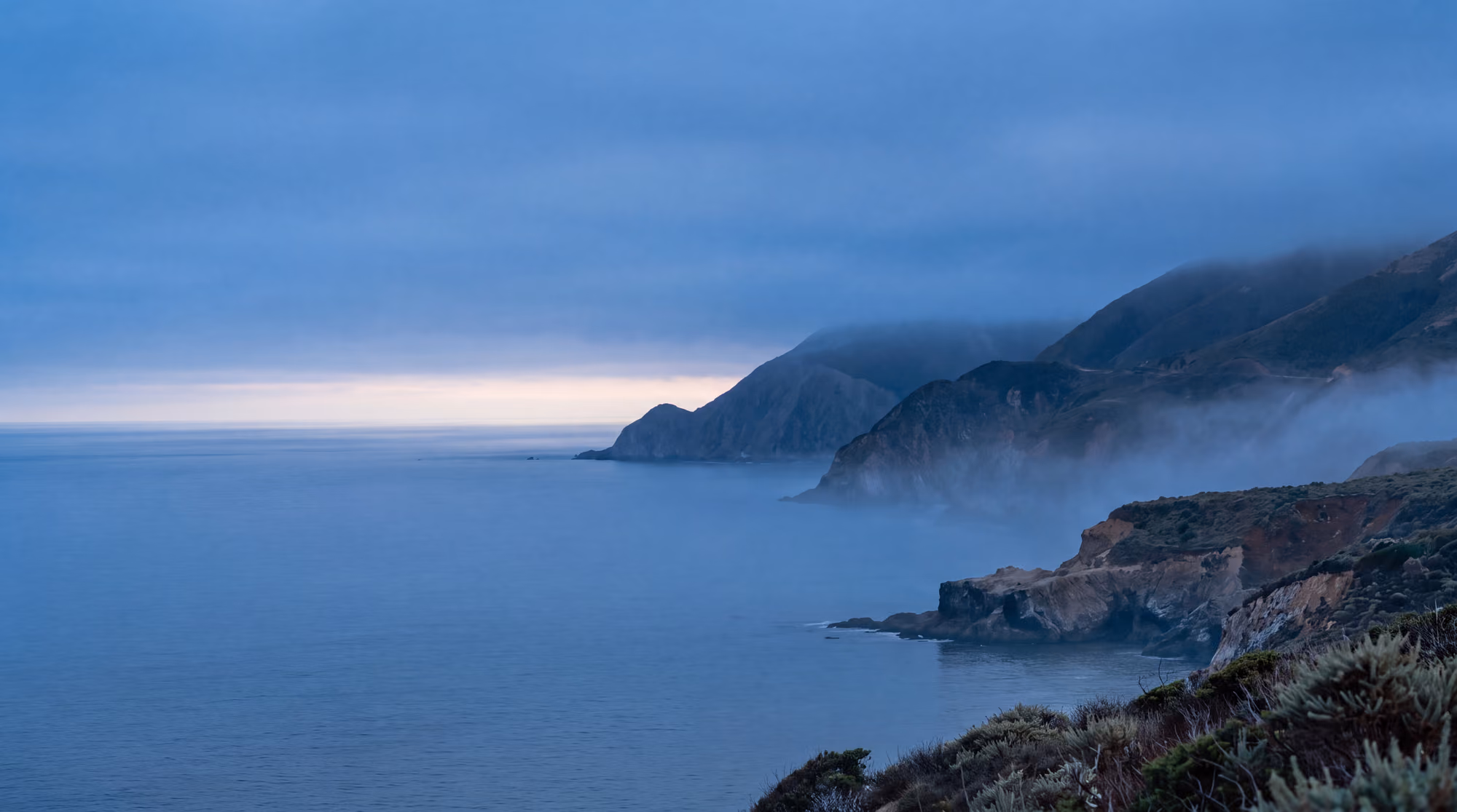 The same headlands and calm coast at blue hour, mist in the folds of the hills